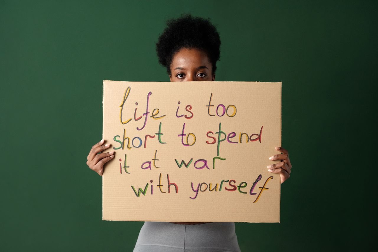 A woman holding a motivational sign with a message about self-acceptance, against a green background.