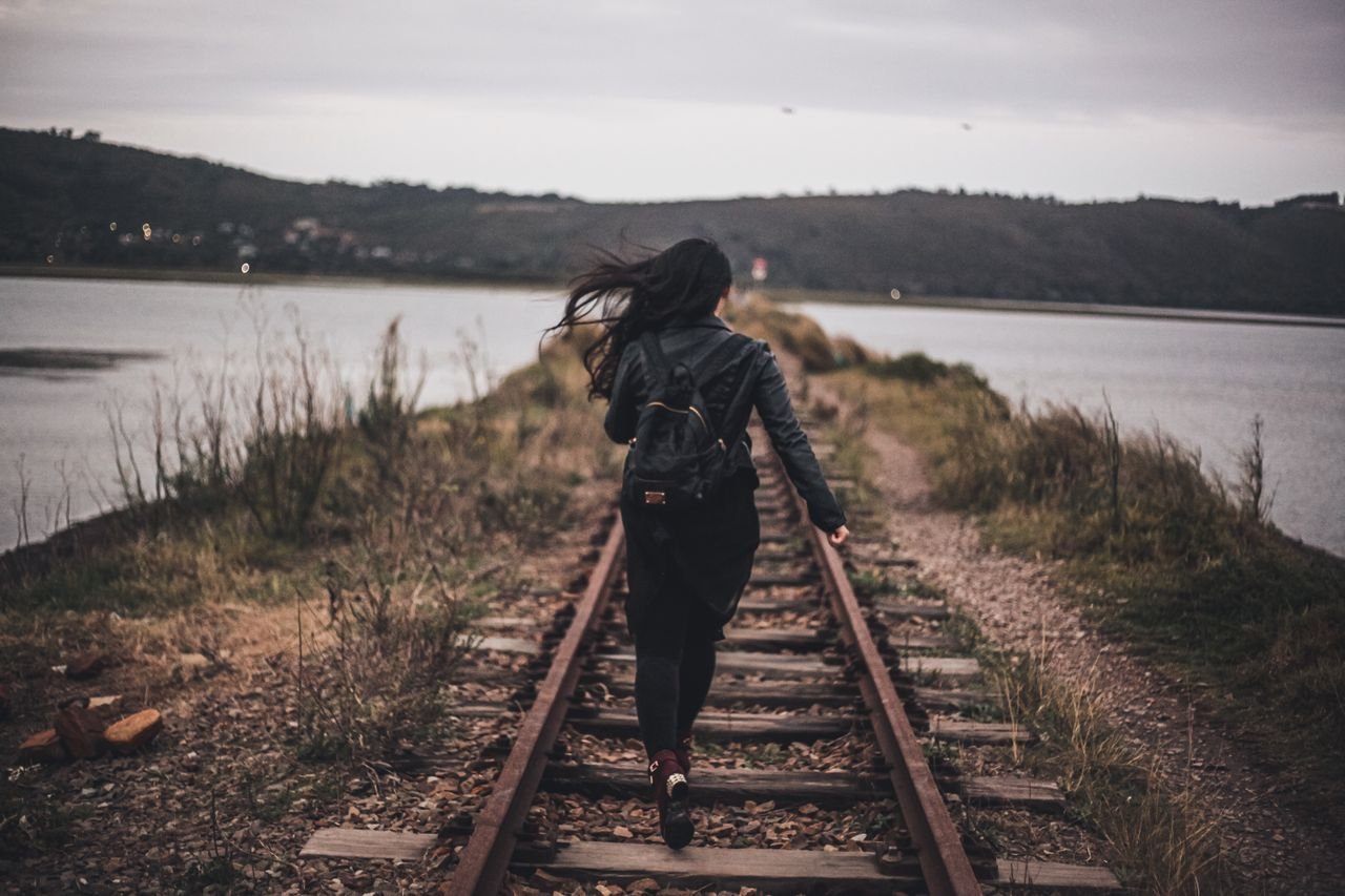 A woman walks, not feeling behind others in life, on railroad tracks next to a serene lake under a cloudy sky, capturing a sense of adventure and solitude.