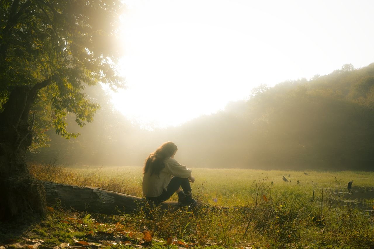 A woman sits on a fallen tree in a misty forest, bathed in warm sunlight. She represents me when I'm enjoying my own company.