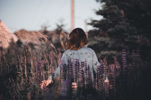 A woman in a denim jacket standing amidst a field of purple lupine flowers at sunset, symbolizing my reflection on my growth.